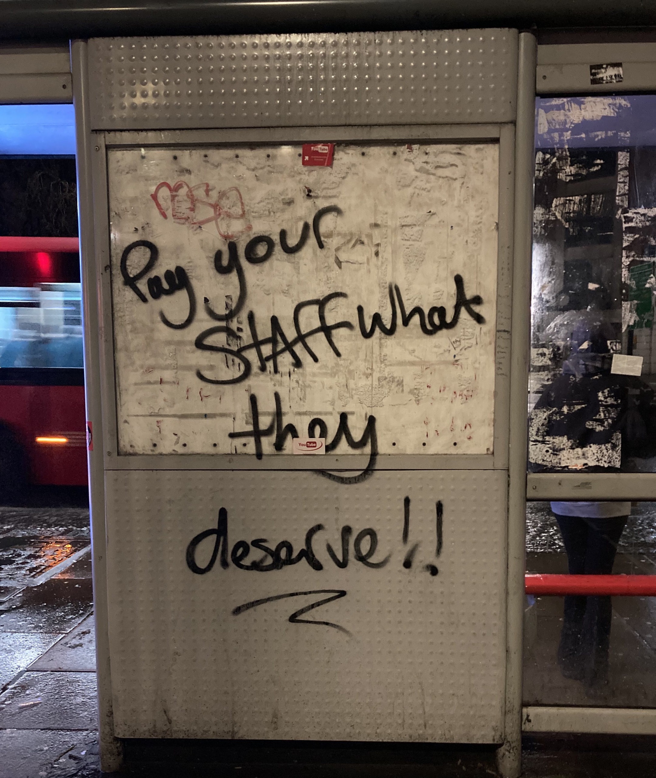A photo of graffiti on a bus stop reading "Pay your STAFF what they deserve!!"