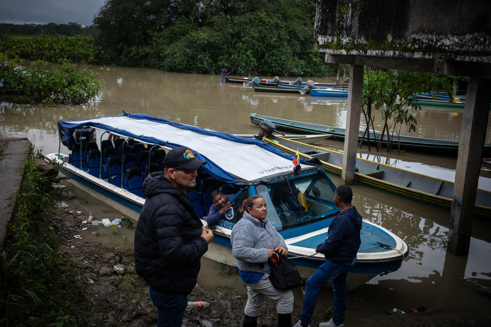 People stand in front of a small boat that is docked on a river.