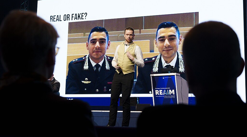 REAIM speaker stands in front of an image that reads "Real or fake?"