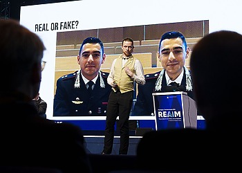 REAIM speaker stands in front of an image that reads "Real or fake?"