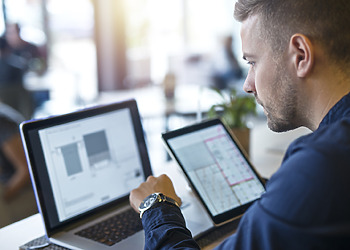Team member of a logistics software development services company looking at his screen with a tablet in his right hand and typing with his left hand.