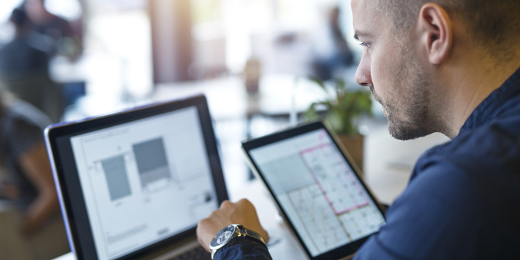 Team member of a logistics software development services company looking at his screen with a tablet in his right hand and typing with his left hand.