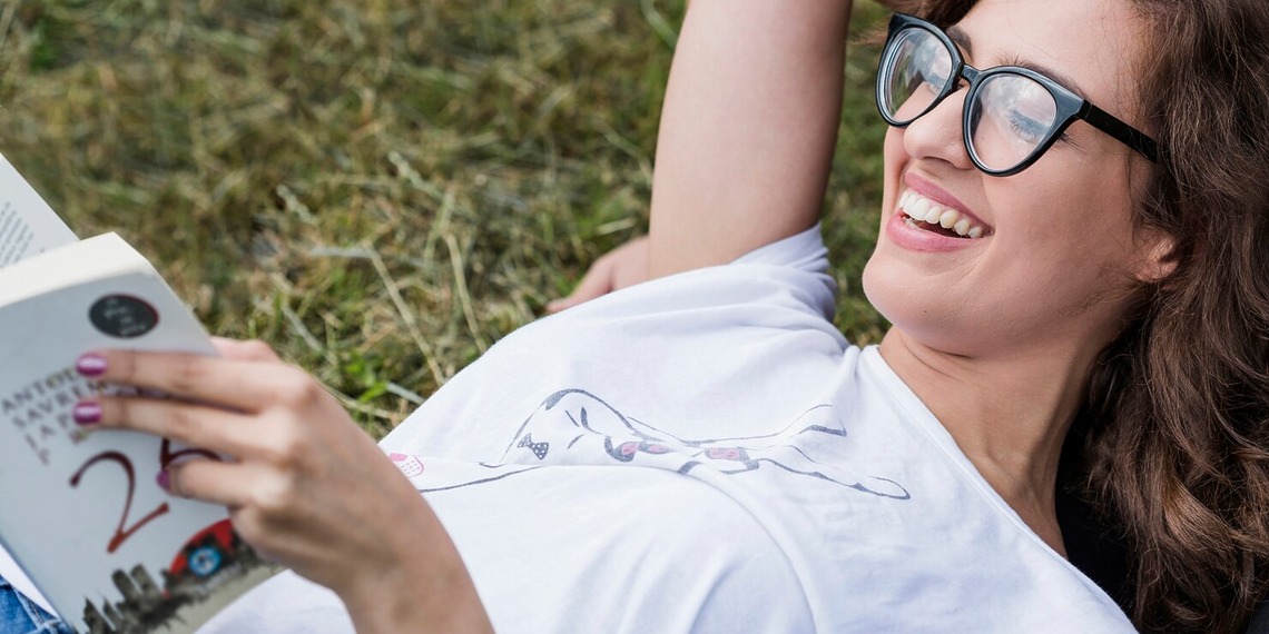A woman laying down on the grass wearing Eco-Friendly Glasses while reading a book.