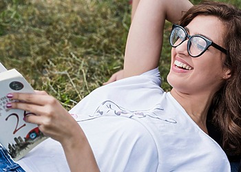 A woman laying down on the grass wearing Eco-Friendly Glasses while reading a book.