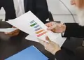 An insurance advisor handing a printed color financial performance bar chart report to a customer during a meeting.