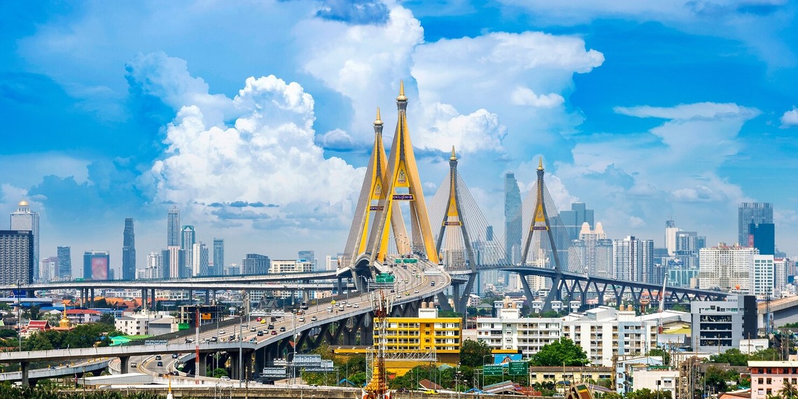 Bhumibol Bridge spanning the Chao Phraya River with the Bangkok city skyline and highway traffic under a blue sky.