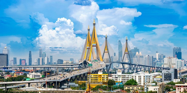 Bhumibol Bridge spanning the Chao Phraya River with the Bangkok city skyline and highway traffic under a blue sky.