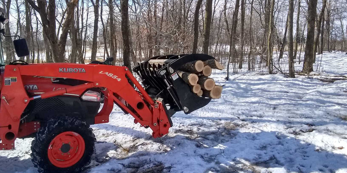 An orange Kubota utility tractor using a heavy duty grapple attachment to carry cut logs over patchy snow in a wooded area.