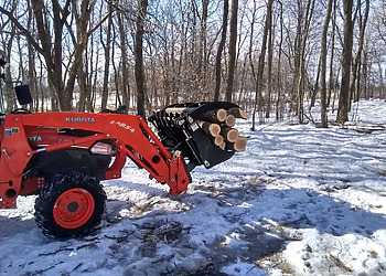An orange Kubota utility tractor using a heavy duty grapple attachment to carry cut logs over patchy snow in a wooded area.