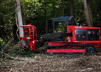 A red tracked forestry tractor using a hydraulic tree shear attachment to selectively cut a tree trunk in dense woods.