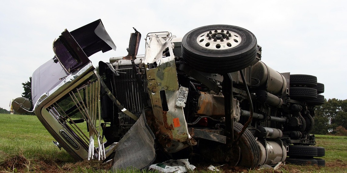 A white commercial semi-truck with extensive front-end cab damage resting on the shoulder of a highway after a major accident.