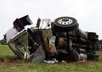 A white commercial semi-truck with extensive front-end cab damage resting on the shoulder of a highway after a major accident.