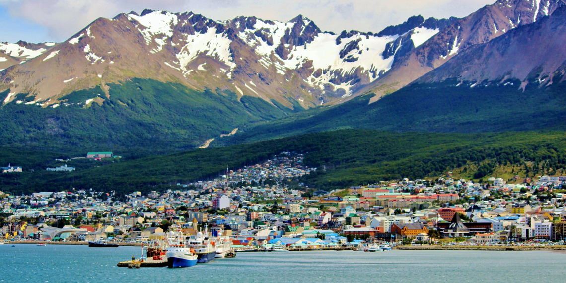 Colorful houses along the waterfront of Ushuaia with snow-capped Martial Mountains rising in the background beneath a blue sky.