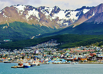 Colorful houses along the waterfront of Ushuaia with snow-capped Martial Mountains rising in the background beneath a blue sky.