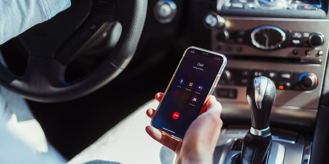 A driver holding a mobile phone showing an active phone call screen while sitting behind the steering wheel inside a car.