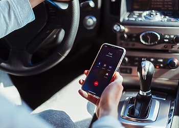 A driver holding a mobile phone showing an active phone call screen while sitting behind the steering wheel inside a car.