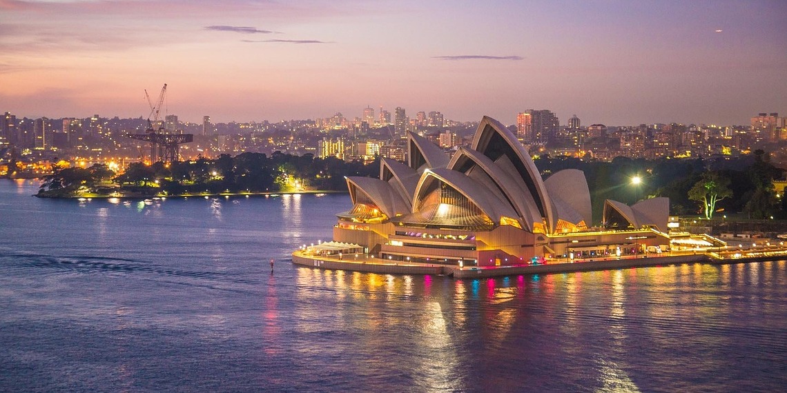 The Sydney Opera House illuminated at dusk along the harbor water with the city skyline in the background.