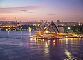 The Sydney Opera House illuminated at dusk along the harbor water with the city skyline in the background.