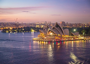 The Sydney Opera House illuminated at dusk along the harbor water with the city skyline in the background.