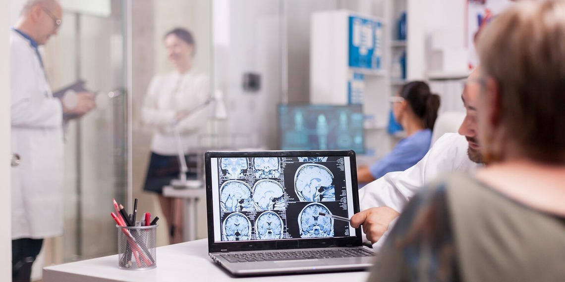 A medical doctor pointing at brain CT scan images on a laptop screen during a patient consultation in a clinic office.