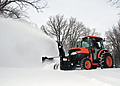 An orange Kubota utility tractor operating an Erskine front-mount snowblower attachment to clear deep snow from a rural driveway.