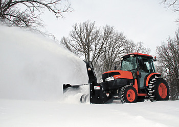 An orange Kubota utility tractor operating an Erskine front-mount snowblower attachment to clear deep snow from a rural driveway.