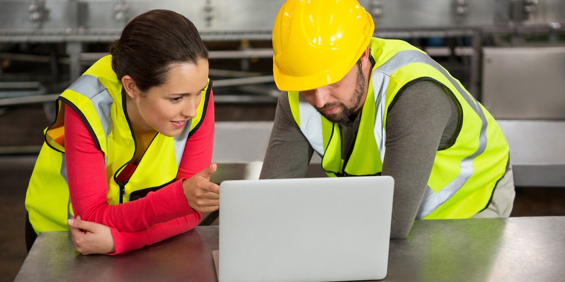 A woman and a man discussing in front of a laptop while wearing safety gear.
