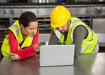 A woman and a man discussing in front of a laptop while wearing safety gear.