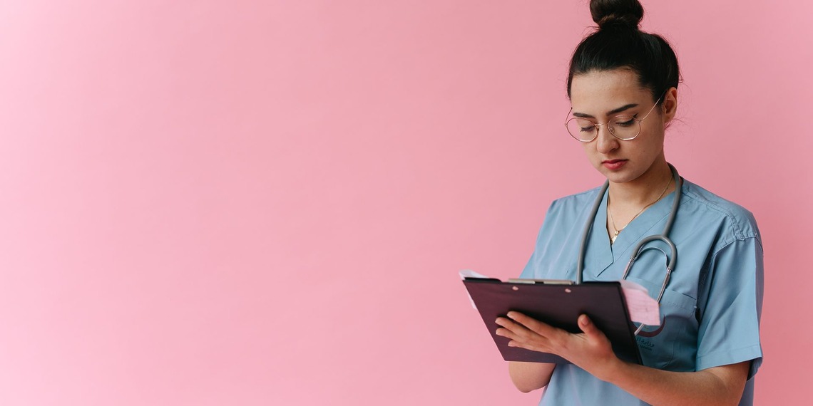 Professional nurse reviewing patient notes on clipboard with pink background, in studio setup.