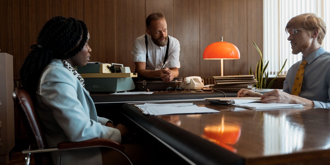 Three colleagues engage in a discussion around a vintage desk in a retro office space.