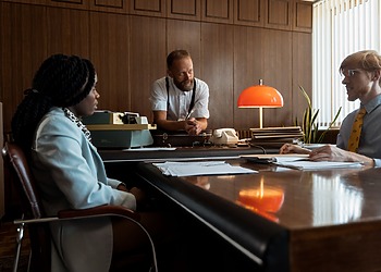 Three colleagues engage in a discussion around a vintage desk in a retro office space.