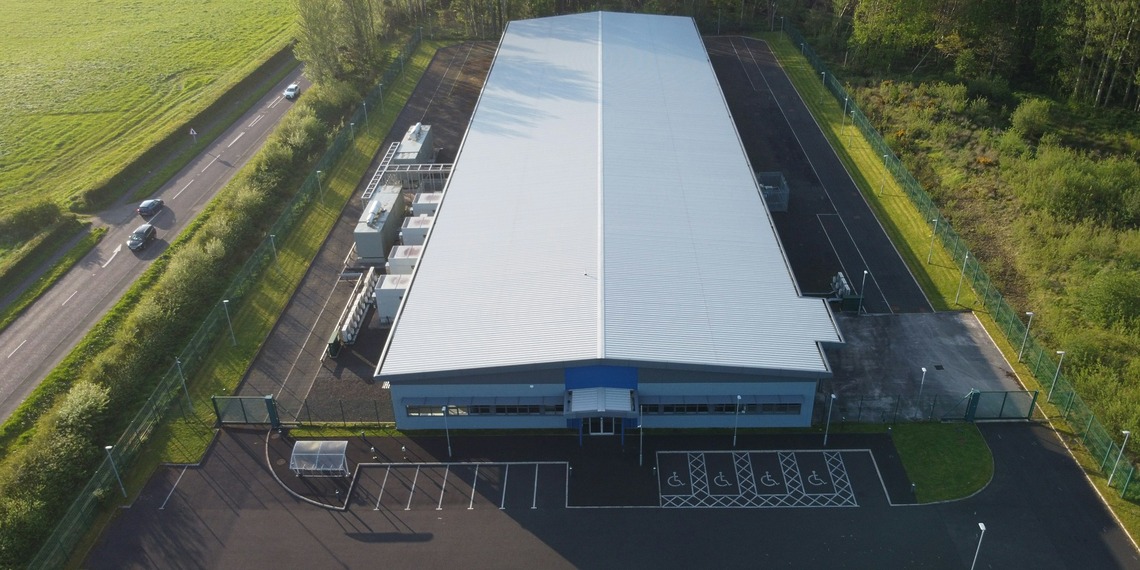 Aerial view of a large white data centre with pitched roof next to a road.