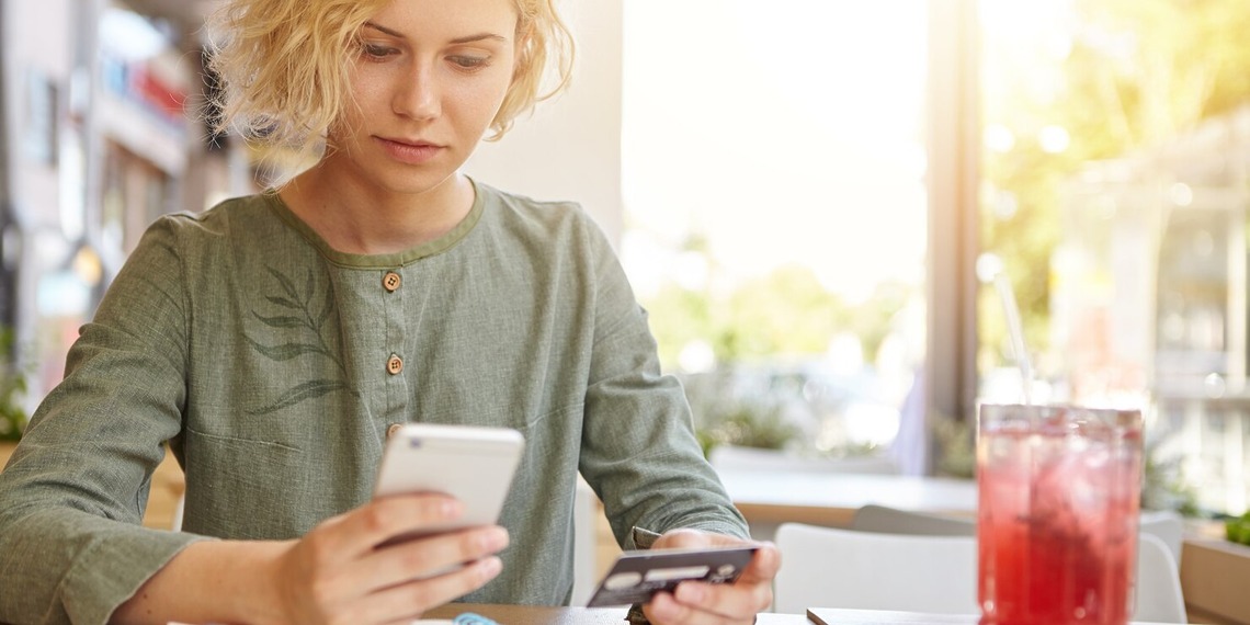 woman holding a phone and her bank card while sitting at a cafè table, drinking a juice.