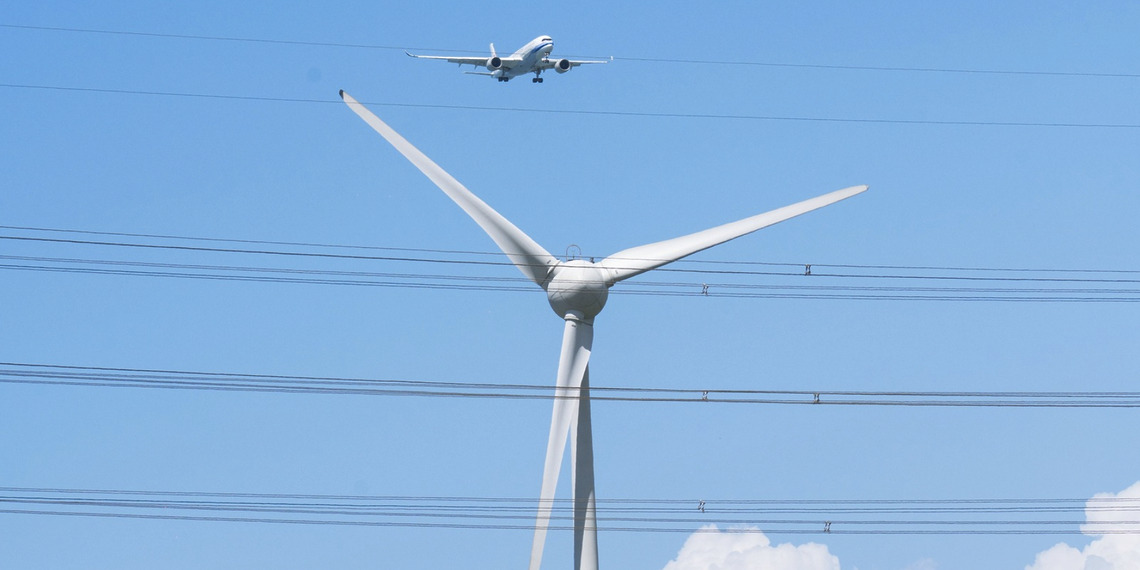 A plane flies over a large white wind turbine.