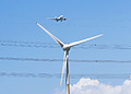 A plane flies over a large white wind turbine.