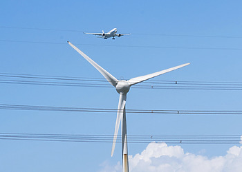 A plane flies over a large white wind turbine.