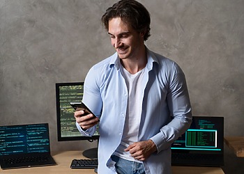 Software developer holding a smartphone in front of multiple coding screens