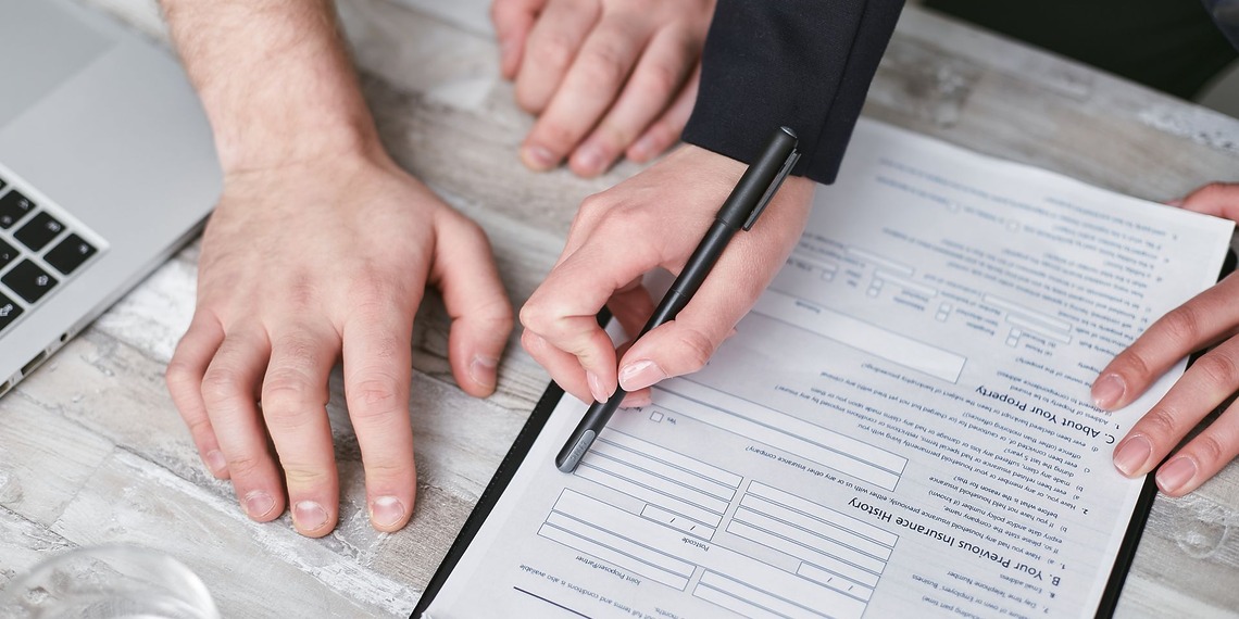 Close-up image of two people signing an insurance policy document on a wooden desk.