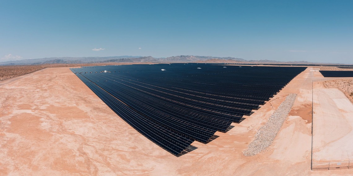 Rows of dark solar panels cover a flat desert plain under a clear blue sky.