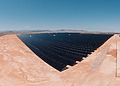 Rows of dark solar panels cover a flat desert plain under a clear blue sky.