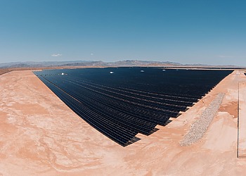 Rows of dark solar panels cover a flat desert plain under a clear blue sky.