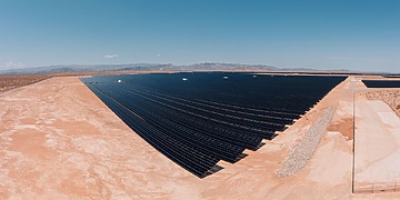 Rows of dark solar panels cover a flat desert plain under a clear blue sky.