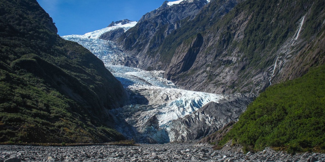 Blue-white glacier ice flows down a narrow valley between steep green slopes under a clear sky.