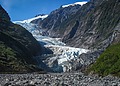 Blue-white glacier ice flows down a narrow valley between steep green slopes under a clear sky.