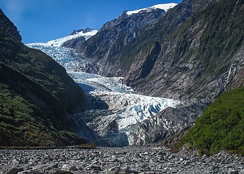 Blue-white glacier ice flows down a narrow valley between steep green slopes under a clear sky.