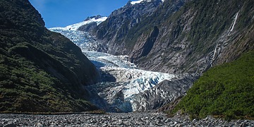 Blue-white glacier ice flows down a narrow valley between steep green slopes under a clear sky.