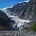 Blue-white glacier ice flows down a narrow valley between steep green slopes under a clear sky.