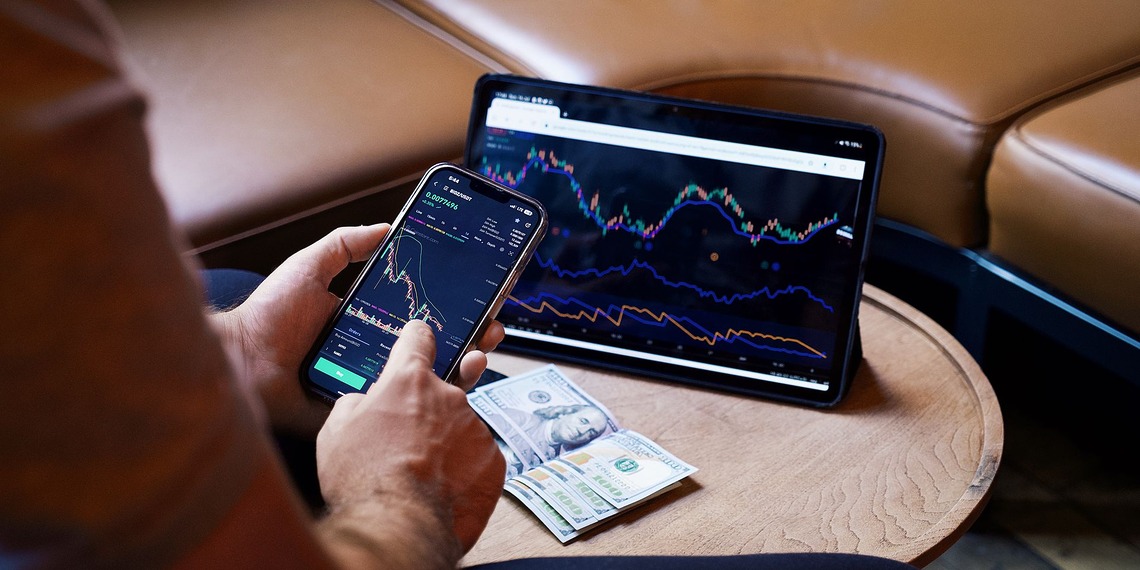 A person using a smartphone and tablet for stock market analysis, with graphs and cash visible.