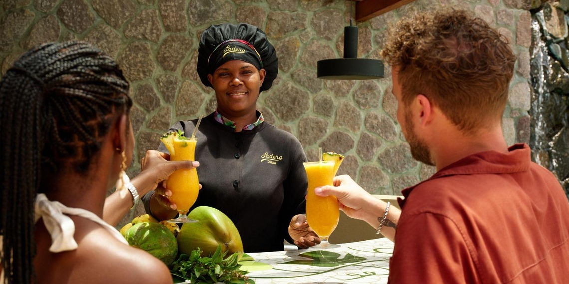A Ladera Resort staff member serves tropical drinks to two guests at an outdoor counter.