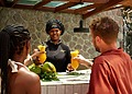 A Ladera Resort staff member serves tropical drinks to two guests at an outdoor counter.